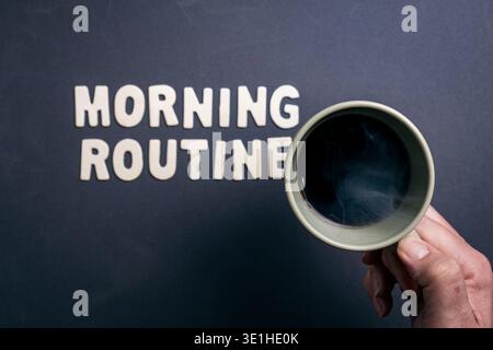 Ein Blick von oben auf eine dampfende Tasse schwarzen Kaffees, die neben hölzernen Buchstaben steht, die „MORGENROUTINE“ auf einer dunklen strukturierten Oberfläche darstellen Stockfoto