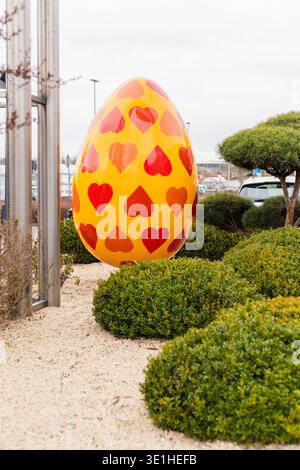 Ein großes gelbes Osterei mit roten Herzformen wird in einem grünen Garten in der Nähe von kleinen Büschen platziert. Es ist tagsüber und im Hintergrund sind Käufer zu sehen Stockfoto
