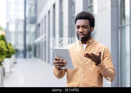 Junger, professioneller Mann, der draußen steht und Frustration und Verwirrung ausdrückt, während er ein digitales Tablet ansieht und sich mit Gesten in Richtung eines digitalen Tablets bewegt und möglicherweise mit technischen Problemen konfrontiert sieht Stockfoto