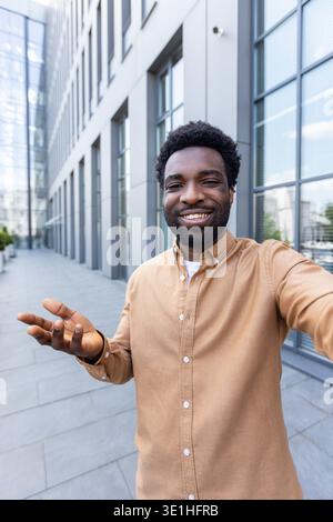Glücklicher junger Mann in einem beigefarbenen Hemd, der vor der Kamera lächelt, während er ein Selfie-Videogespräch vor einem modernen Bürogebäude nimmt und gute Nachrichten mit Freunden oder Kollegen teilt Stockfoto