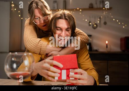 Verliebte Paare feiern Valentinstag während eines romantischen Abendessens mit der Frau, die ihren Ehemann umarmt und Geschenke in der Küche zu Hause austauscht. Kopierbereich Stockfoto