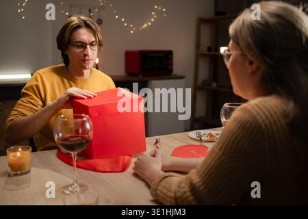 Ein Paar mittleren Alters feiert den Valentinstag zu Hause während eines romantischen Abendessens und tauscht Geschenke aus, während er am Tisch sitzt. Konzept eines romantischen Abendessens, Stockfoto