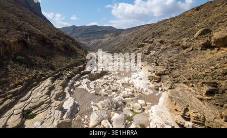 Breiter felsiger Canyon mit trockenem Flussbett, weißen Felsblöcken und mehrschichtigen Steinmauern unter blauem Himmel mit verstreuten Wolken, zerklüftete Wüstenberglandschaft. Ka Stockfoto