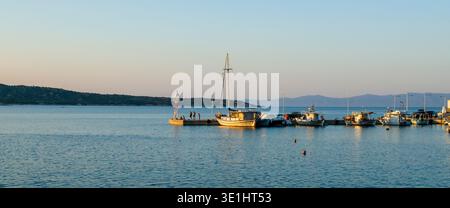 Friedliche Hafenszene mit angelegten Fischerbooten, einem rot-weißen Leuchtturm und Bergen im Hintergrund während der goldenen Stunde. Das ruhige Wasser Stockfoto