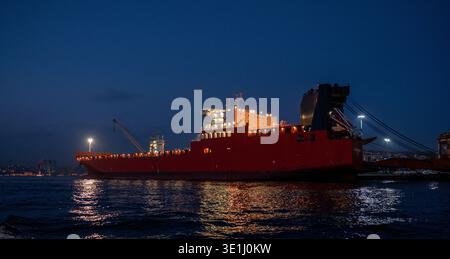 Ein riesiges rotes Frachtschiff, beleuchtet von künstlichen Lichtern, das während der Dämmerung an einem Industriehafen angedockt ist. Die helle Beleuchtung des Schiffes reflektiert Schönheit Stockfoto