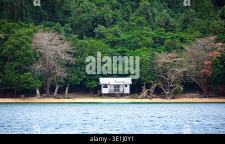 Ein kleines weißes Haus auf Stelzen liegt an einem Sandstrand, der von einem dichten grünen Wald umgeben ist. Das Haus hat eine Veranda mit Stühlen und eine Treppe, die dow führt Stockfoto