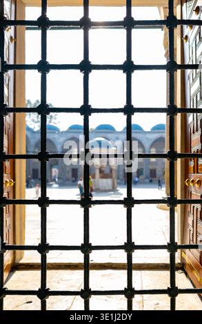 Ein Blick durch dekorative schmiedeeiserne Fensterstangen zeigt einen sonnendurchfluteten historischen europäischen Innenhof mit klassischer Bogenarchitektur und Spaziergänge Stockfoto