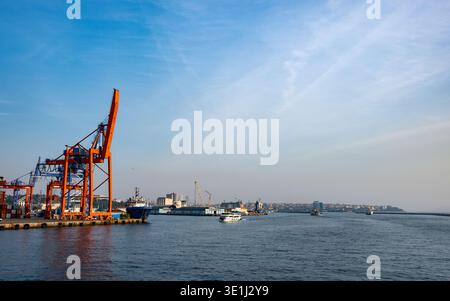Industriehafenanlage mit großen orangen Containerkränen entlang der Uferpromenade mit Frachtschiffen und Schiffen im Hafen. Klarer blauer Himmel Stockfoto
