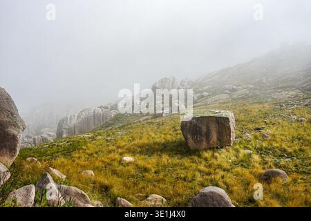Großer Felsbrocken auf einem grasbewachsenen Hügel mit verstreuten Felsen und Nebel, der die Berglandschaft bedeckt Stockfoto