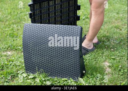 Ein schwarzer Kunststoffstuhl für den Außenbereich ist teilweise auf einem grasbewachsenen Rasen montiert. Die Beine einer Person sind im Hintergrund sichtbar. Stockfoto
