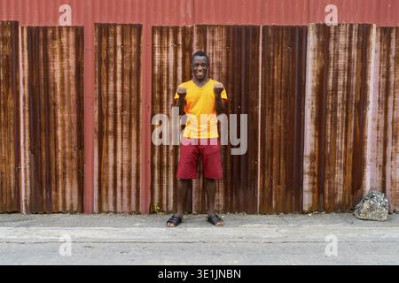 Junge glücklich schwarzen afrikanischen Mann lächelnd und im Stehen gegen die alten rostigen Blech Wand suchen begeistert Stockfoto