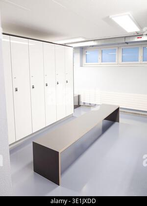 Moderner Umkleideraum mit weißen Schließfächern, einer langen Holzbank und großen Fenstern, die Tageslicht bieten, schaffen eine saubere und funktionale Umgebung Stockfoto