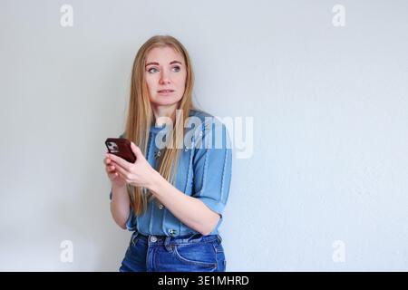 Junge Frau, die ein Smartphone hält und mit überraschtem Ausdruck beiseite schaut. Mobilkommunikation und Technologiekonzept. Kopierbereich Stockfoto