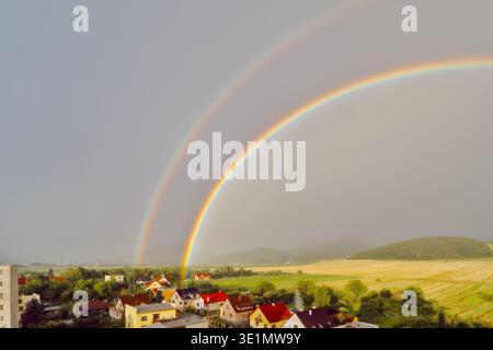 Eine weite Aufnahme, die einen lebhaften doppelten Regenbogenbogen zeigt, der sich über eine ländliche Stadt mit roten Häusern, grünen Feldern und fernen Hügeln erstreckt. Stockfoto