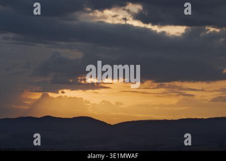 Eine stimmungsvolle abendliche Landschaft mit orangefarbenem Sonnenuntergang, das durch dunkelgraue Wolken über die Silhouetten der sanften Hügel blickt. Stockfoto