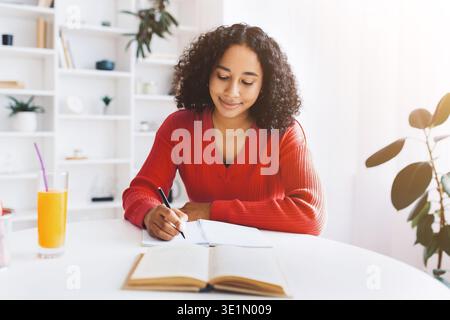 Die junge Frau schreibt in Notizbuch, während sie im hellen Raum mit Pflanzen trinkt Stockfoto