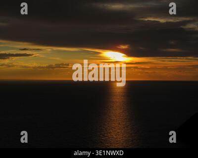 Ein dramatischer goldener Sonnenuntergang über einem ruhigen Meer, mit warmem Licht, das durch dunkle Wolken bricht und sich über das Wasser reflektiert. Stockfoto