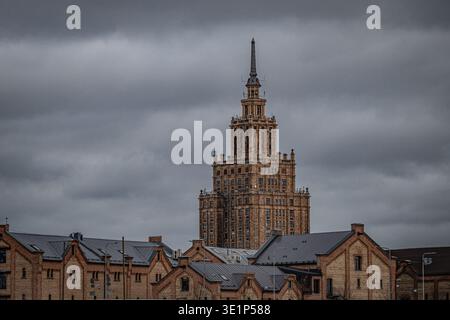 Das Gebäude der lettischen Akademie der Wissenschaften (Stalinistisches Wolkenkratzer aus der Sowjetzeit) erhebt sich über den Lagerhäusern am Flussufer unter dramatischem Himmel, Riga, Lettland Stockfoto