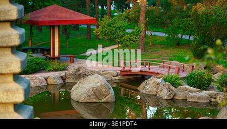 Eine ruhige Parkszene mit einer roten Pagode, einer bezaubernden Holzbrücke, die sich über einen Teich erhebt, und üppigem Grün, das sich im stillen Wasser spiegelt Stockfoto