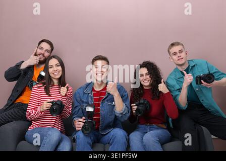Gruppe von Fotografen mit professionellen Kameras, die Daumen hoch auf dem Sofa vor farbigem Hintergrund zeigen Stockfoto
