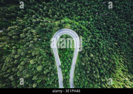 Atemberaubendes Luftbild der Drohne, das eine dramatische Haarnadelkurve auf einer gewundenen Bergstraße, umgeben von dicken Nadelwäldern, aufnimmt. Die Asphaltstraße Stockfoto