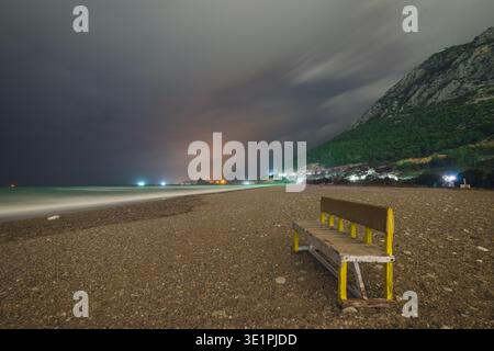 Langzeitaufnahme einer leeren Holzbank auf einem Kieselstrand. Dunkler, stimmungsvoller Himmel, beleuchtete ferne Küste und sanfte Wellen des Meeres im Hintergras Stockfoto