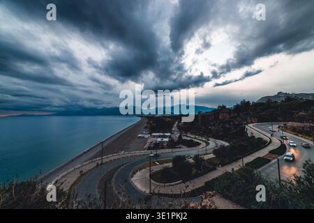Ein Weitwinkel-Panoramablick auf die Küste bei einem stürmischen Sonnenuntergang. Gewundene Küstenstraßen mit Autolichtern, ein langer Kiesstrand, das Mittelmeer Stockfoto