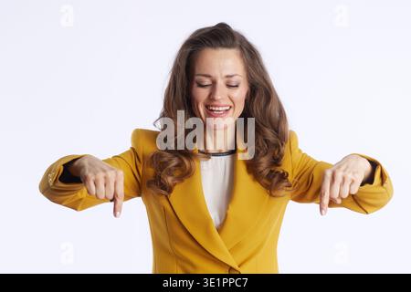Eine glückselige Geschäftsfrau, lächelt mit geschlossenen Augen und zeigt mit beiden Zeigefingern nach unten. In einem leuchtenden Senfblazer gekleidet, vermittelt sie fröhliche gui Stockfoto