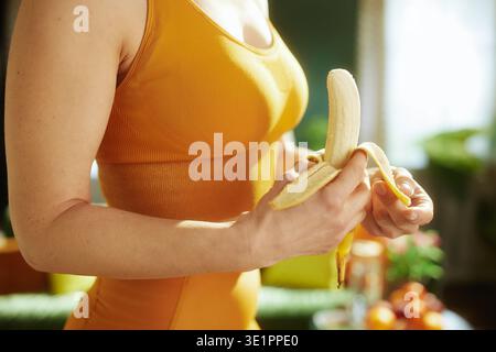 Eine Frau in orangefarbener Sportausrüstung schält eine reife Banane in einem hellen Innenbereich und konzentriert sich dabei auf sauberes Essen und Energie für ihre Fitnessroutine. Stockfoto