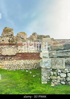 Eine Steinmauer, die Teil historischer Ruinen ist. Die Wand zeigt Grasflecken vorne und einen blauen Himmel darüber. Stockfoto