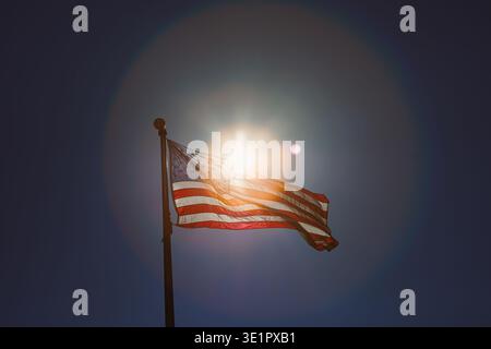 Die Flagge der Vereinigten Staaten flattert auf einem Pfosten unter der hellen Sonne. Sonnenstrahlen leuchten um die Flagge und bilden einen auffälligen Umriss gegen den Kleinen Stockfoto