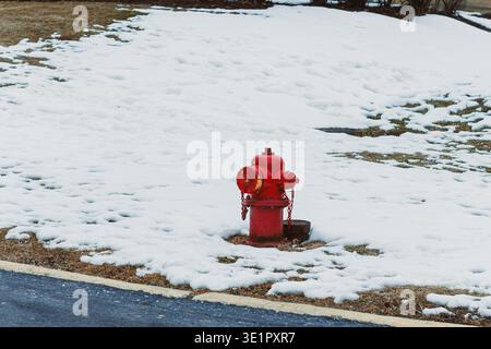 Im Winter ist ein roter Hydrant auf einem schneebedeckten Gras neben einer schwarzen asphaltierten Straße in einem Wohnviertel zu sehen. Stockfoto