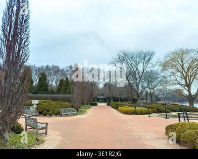 Ein Pfad führt durch einen Park mit Bänken auf jeder Seite. Entlang des Weges stehen lautlose Bäume. Der Himmel ist bewölkt, und das Gebiet ist nahe an einem Wasserkörper. Stockfoto