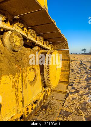 Gelbe Baggerspuren ruhen auf sandigem Boden auf einer Baustelle in der Nähe des Strandes. Die Sonne geht im Hintergrund unter und die Menschen bewegen sich in der Ferne Stockfoto