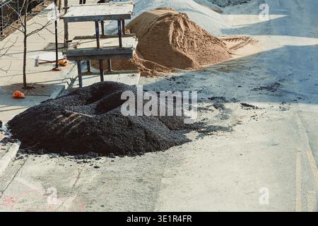 Arbeiter sind am Straßenbau auf einer Stadtstraße beteiligt. Neben Gerüsten befinden sich große Anhäufungen aus schwarzem Asphalt und Sand. In der Gegend ist viel los Stockfoto
