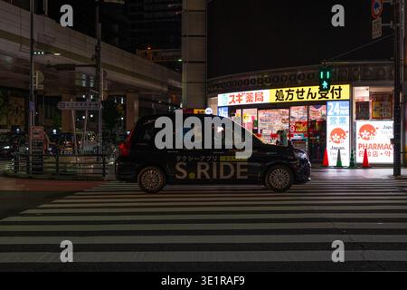 Ein schwarzes Taxi fährt spät in der Nacht über eine Kreuzung vor einer hell beleuchteten Apotheke in Roppongi, Tokio, Japan. Stockfoto