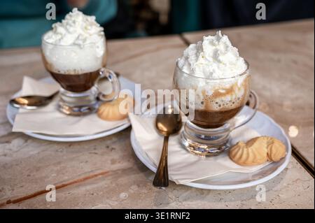 Zwei klare Gläser gefüllt mit mehrschichtigem Kaffee und großzügig mit flauschiger Schlagsahne, serviert mit kleinen Keksen auf Untertassen und Löffeln auf einem mar Stockfoto