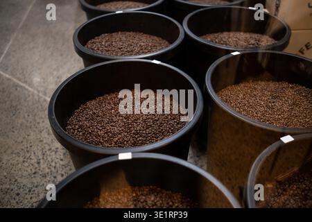 Große schwarze Behälter gefüllt mit gerösteten Kaffeebohnen, die in einem Lagergebäude auf einem Betonboden mit sichtbaren Etiketten angeordnet sind Stockfoto
