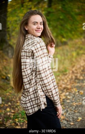 Eine junge Frau steht auf einem Weg, bedeckt mit gefallenen Blättern und trägt ein kariertes Hemd. Sie lächelt und genießt die wunderschöne Herbstlandschaft, die sie umgibt. Stockfoto