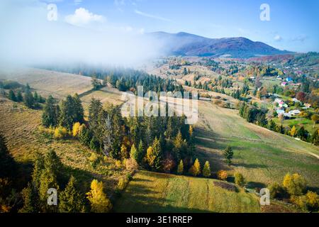 Malerisches Dorf eingebettet in sanfte Hügel, in Herbstfarben mit grünen und gelben Feldern getaucht. Nebelschwaden ziehen durch die Landschaft und verleihen der ruhigen ländlichen Szene eine traumhafte Atmosphäre. Stockfoto