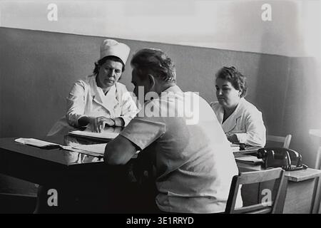 Ein Schwarzweiß-Archivfoto aus dem Jahr 1977 zeigt eine medizinische Beratung im Krankenhaus der AIZ-Fabrik in der Stadt Slawjansk. Eine Ärztin und eine Krankenschwester in weißen Mänteln sitzen an einem Schreibtisch, untersuchen sorgfältig Dokumente und sprechen mit einem männlichen Patienten, der ihnen gegenüber sitzt. Die Szene vermittelt die ernsthafte und gründliche Herangehensweise des sowjetischen medizinischen Systems an die Arbeitsmedizin. Dieses historische Bild erfasst Routine-klinische Praxis, typische Krankenhauseinrichtungen und den friedlichen Donbass vor dem Krieg. Stockfoto