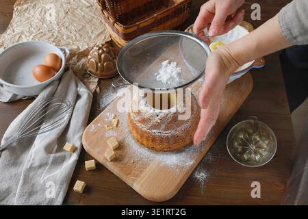 Nahaufnahme von weiblichen Händen, die Biskuitkuchen auf hölzernem Schneidebrett stauben. Gemütliche Küche mit Eiern, Schneebesen, Backformen und Zuckerwürfeln im Dunkeln Stockfoto