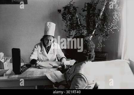 Ein Schwarzweiß-Archivfoto aus dem Jahr 1977 zeigt eine Ärztin in einem weißen Mantel und einer hohen Kappe, die eine medizinische Beratung mit einem Patienten im AIZ-Fabrikkrankenhaus in der Stadt Slawjansk durchführt. Die Ärztin überprüft sorgfältig die medizinischen Aufzeichnungen an ihrem Schreibtisch, der mit einem Vintage-Blutdruckmessgerät ausgestattet ist. Dieses historische Bild bewahrt perfekt die übliche klinische Praxis, die Arbeitsmedizin, den routinemäßigen Krankenhausablauf und die Realität des friedlichen Donbas vor dem Krieg. Stockfoto
