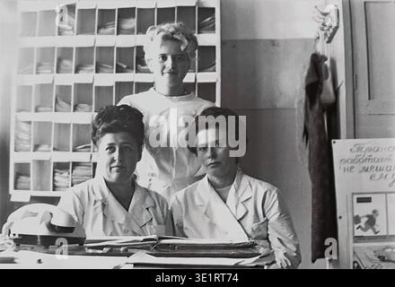 Ein Schwarzweiß-Archivfoto, das 1977 aufgenommen wurde, zeigt ein posiertes Gruppenporträt von drei Ärztinnen am belebten Registerschalter der Klinik des AIZ-Fabrikkrankenhauses in der Stadt Slawjansk. Zwei Frauen in weißen Mänteln sitzen, während eine andere hinter ihnen vor einem Hintergrund von weitläufigen Holzregalen steht, in denen Patientenakten aufbewahrt werden. Dieser historische Schnappschuss bewahrt die Gesichter sowjetischer medizinischer Fachkräfte, den Arbeitsalltag und den friedlichen Donbass vor dem Krieg. Stockfoto