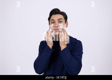Junger Mann, der Feuchtigkeitscreme auf das Gesicht aufträgt und sieht Stockfoto