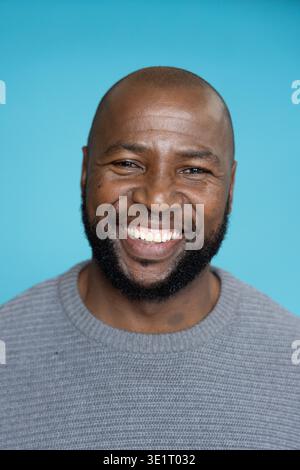 Afroamerikaner mittleren Alters lächelt und trägt einen grauen Pullover im Studio mit türkisfarbenem Hintergrund Stockfoto