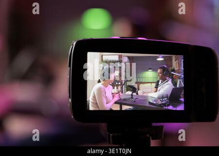 Nahaufnahme mit Fokus auf Vorschaubild auf einer professionellen Kamera mit Podcast-Moderator und afroamerikanischem Gast, der Gespräche im unscharfen Hintergrund genießt. Professionelle Aufnahmegeräte im Heimstudio Stockfoto