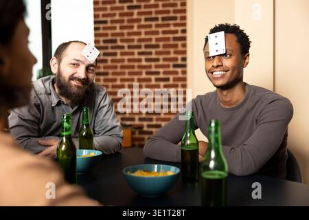 afroamerikanische und kaukasische Männer sitzen mit Karten auf der Stirn, umgeben von Snacks und Getränken während des Treffens. Das Lachen bricht aus, wenn sich Freunde am Tisch versammeln und ein unbeschwertes Ratespiel genießen. Stockfoto