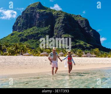 In der atemberaubenden Landschaft von Mauritius spazieren ein Paar Hand in Hand am kristallklaren Wasser entlang. Der lebhafte grüne Berg ragt majestätisch hinter sich, perfekt für einen romantischen Kurzurlaub. Stockfoto
