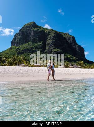 Goldener Sand und klares Wasser schaffen eine ruhige Atmosphäre, während ein Paar einen gemütlichen Spaziergang am Strand von Mauritius genießt, umgeben von beeindruckenden Berglandschaften von Le Morne Mauritius Stockfoto